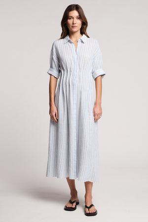 A woman wears the SARON DRESS by Velvet by Graham & Spencer—a light blue and white striped cotton-linen dress with rolled sleeves—paired with black sandals, standing against a plain background.