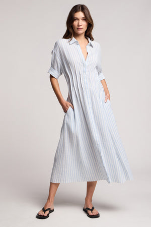 Model wears the SARON DRESS by Velvet by Graham & Spencer, a light blue and white striped cotton-linen dress with rolled sleeves and pockets, paired with black sandals.