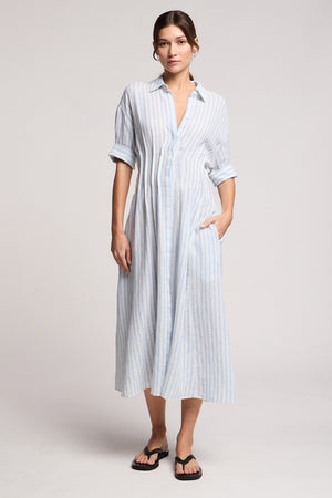 A woman wears the Velvet by Graham & Spencer SARON DRESS—a light blue and white striped, embroidered, button-down cotton-linen dress with a pleated waist, rolled sleeves, pockets—and black flip-flops against a plain background.
