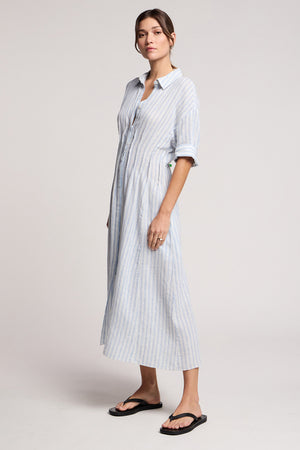 A woman wears the SARON DRESS by Velvet by Graham & Spencer, a light blue and white striped cotton-linen dress with a pleated waist and rolled sleeves, paired with black sandals against a plain background.