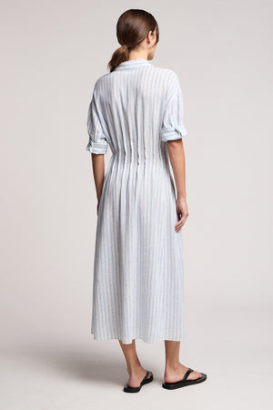 A person stands with their back to the camera, wearing the Velvet by Graham & Spencer SARON DRESS—a light cotton-linen dress with vertical embroidered stripes and rolled-up sleeves—paired with black sandals.