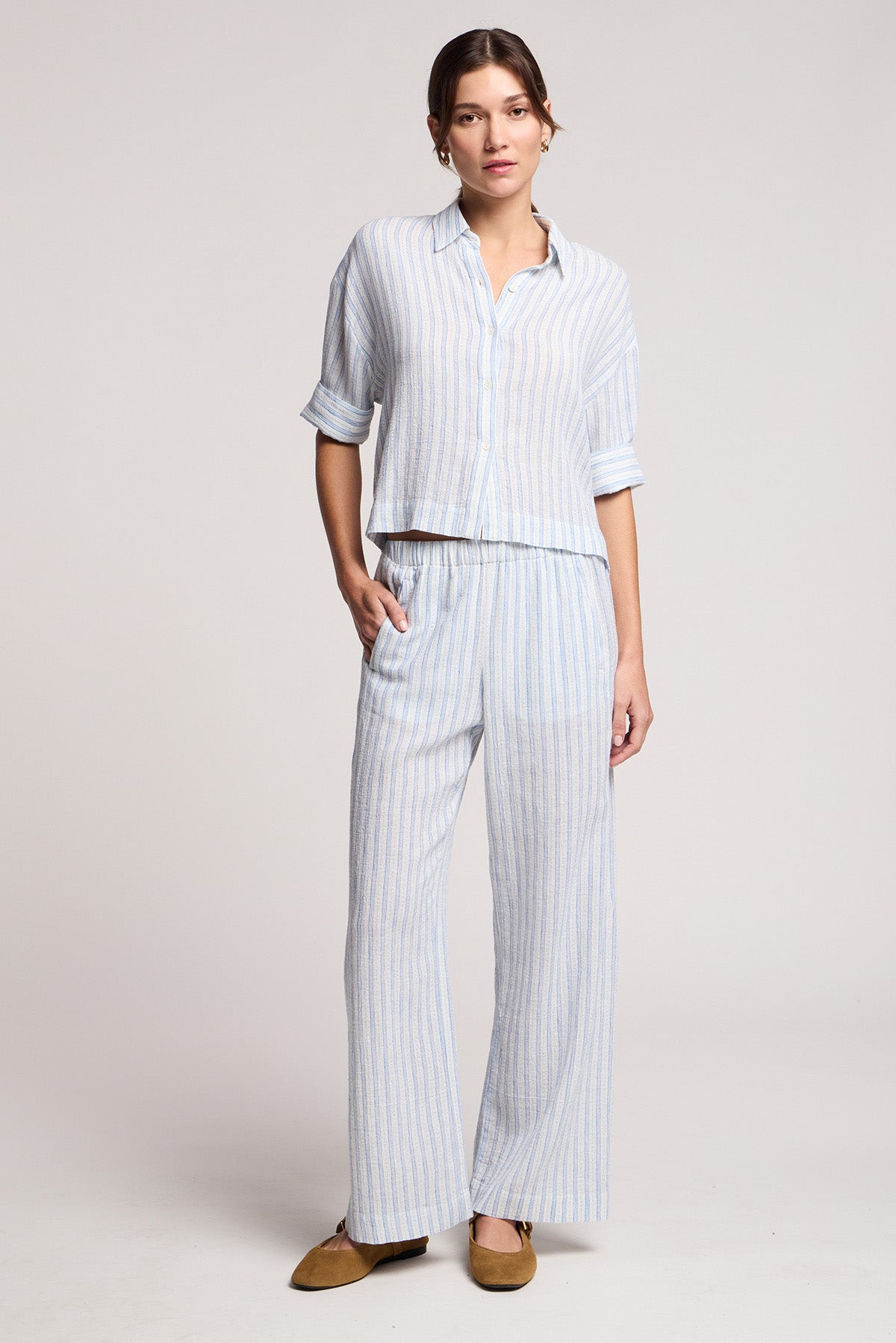 A person wears the EVONY PANT by Velvet by Graham & Spencer, styled in a light blue and white striped cotton-linen blend with a matching button-up shirt and brown flats, standing against a plain background.-41645017956545