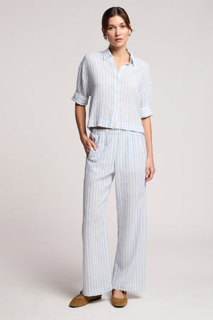 A person wears the EVONY PANT by Velvet by Graham & Spencer, styled in a light blue and white striped cotton-linen blend with a matching button-up shirt and brown flats, standing against a plain background.