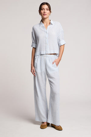 A woman wears the GREYA SHIRT by Velvet by Graham & Spencer, a loose-fitting light blue and white cotton-linen blend summer shirt with embroidered stripes, paired with matching wide-leg pants and brown flats.