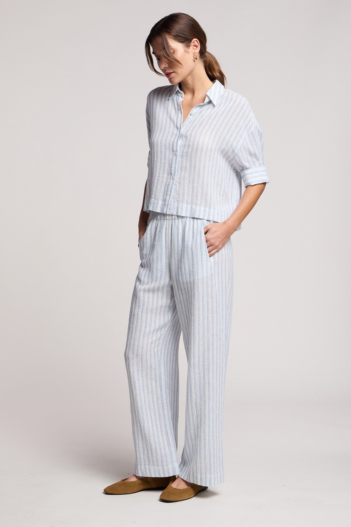 A woman stands in a relaxed pose wearing Velvet by Graham & Spencer’s EVONY PANT—a light blue and white striped, cotton-linen blend wide-leg pant with an elastic waist—styled with a matching shirt and brown flats.