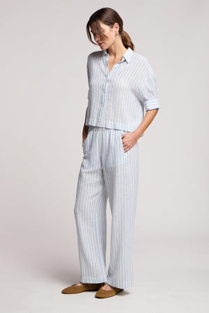 A woman stands in a relaxed pose wearing Velvet by Graham & Spencer’s EVONY PANT—a light blue and white striped, cotton-linen blend wide-leg pant with an elastic waist—styled with a matching shirt and brown flats.