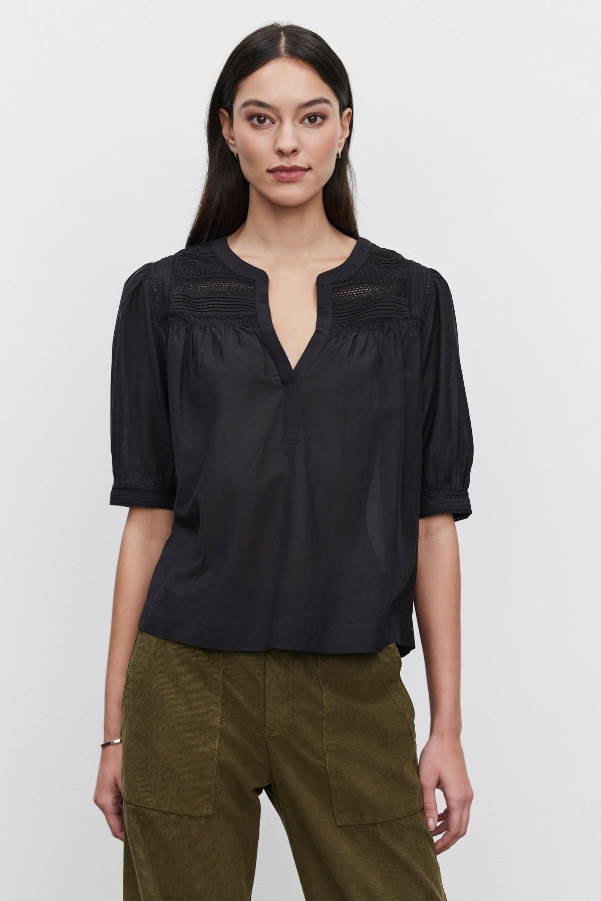   A person with long dark hair wearing the MARCI TOP from Velvet by Graham & Spencer, featuring embroidered lace details, and olive green pants stands against a plain white background. 