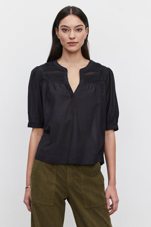 A person with long dark hair wearing the MARCI TOP from Velvet by Graham & Spencer, featuring embroidered lace details, and olive green pants stands against a plain white background.