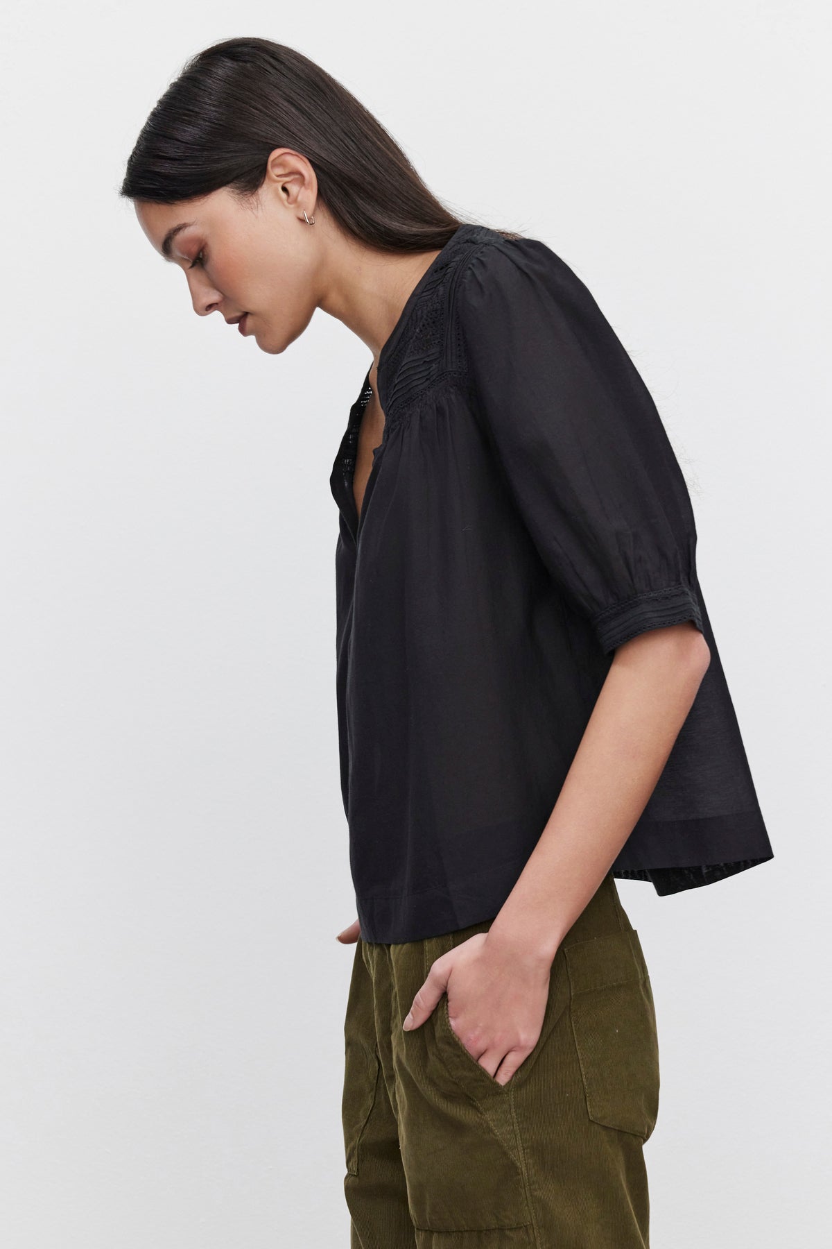   Profile view of a person with long dark hair, wearing the MARCI TOP by Velvet by Graham & Spencer, an embroidered lace detail black blouse with three-quarter sleeves, paired with olive-green pants, standing against a plain white background. 