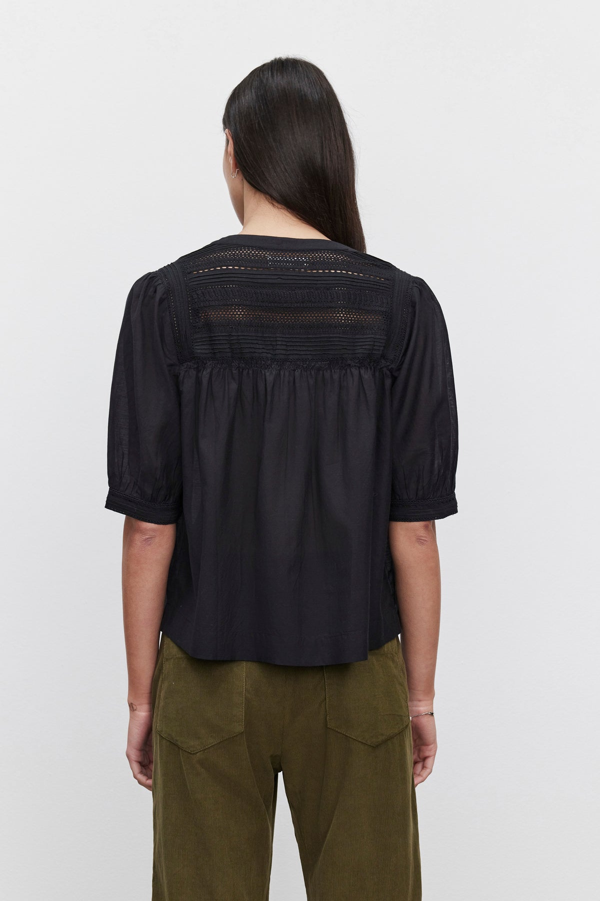   A person with long brown hair is seen from the back wearing the MARCI TOP by Velvet by Graham & Spencer, a black short-sleeved blouse featuring embroidered lace details on the upper back, paired with green pants. 