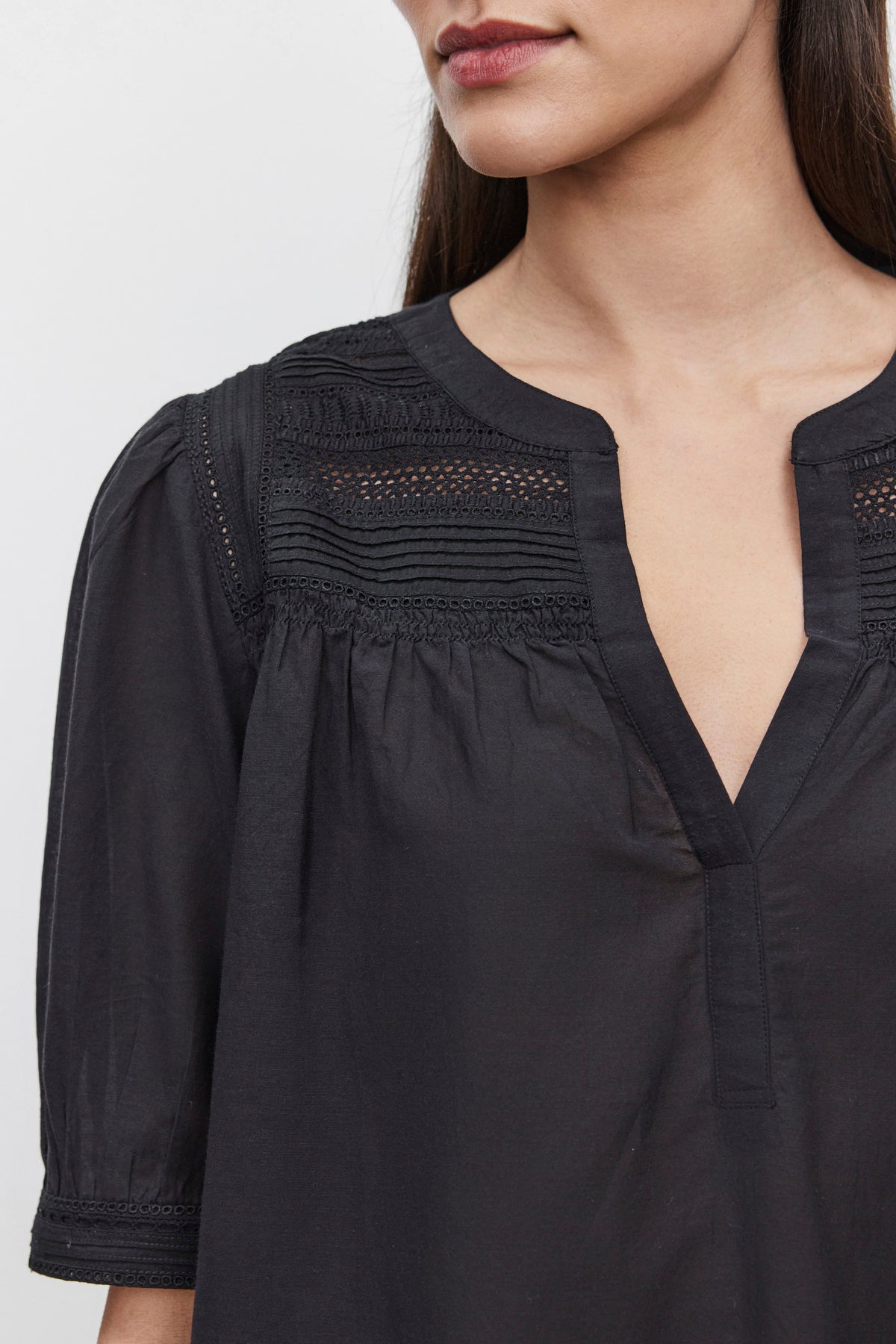   Close-up image of a person wearing the MARCI TOP by Velvet by Graham & Spencer, showcasing its black blouse design with intricate embroidered lace details and a deep neckline. Only the lower part of the face and upper part of the garment are visible, emphasizing the craftsmanship of the cotton blend fabric. 