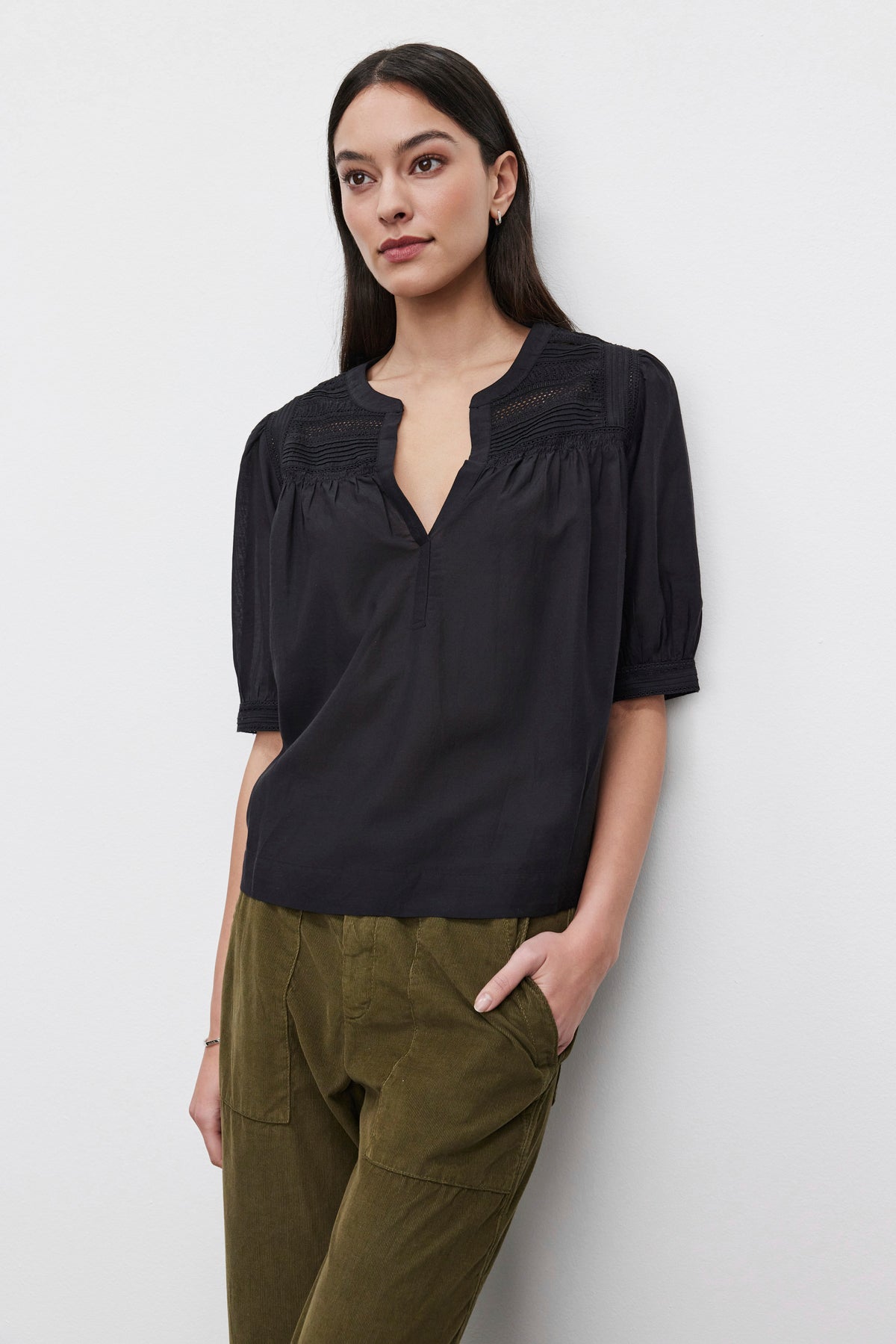   A woman with long dark hair, wearing the MARCI TOP by Velvet by Graham & Spencer — a black blouse showcasing embroidered lace details — and olive green pants, stands against a plain background with one hand in her pocket. 