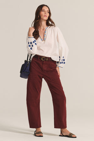 A woman stands in a relaxed pose wearing the AMANI BLOUSE by Velvet by Graham & Spencer, a flowy white cotton top with blue embroidery, paired with maroon pants, black sandals, and a navy handbag.