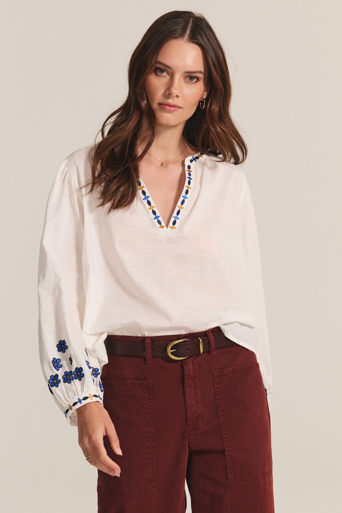 A woman wears the AMANI BLOUSE by Velvet by Graham & Spencer, a white cotton top with blue embroidery, paired with maroon high-waisted pants, looking toward the camera.