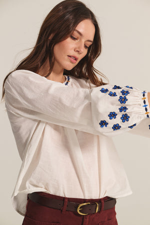 She wears the AMANI BLOUSE by Velvet by Graham & Spencer, a flowy white embroidered top with blue flowers on the sleeves, styled with maroon pants and a brown belt.
