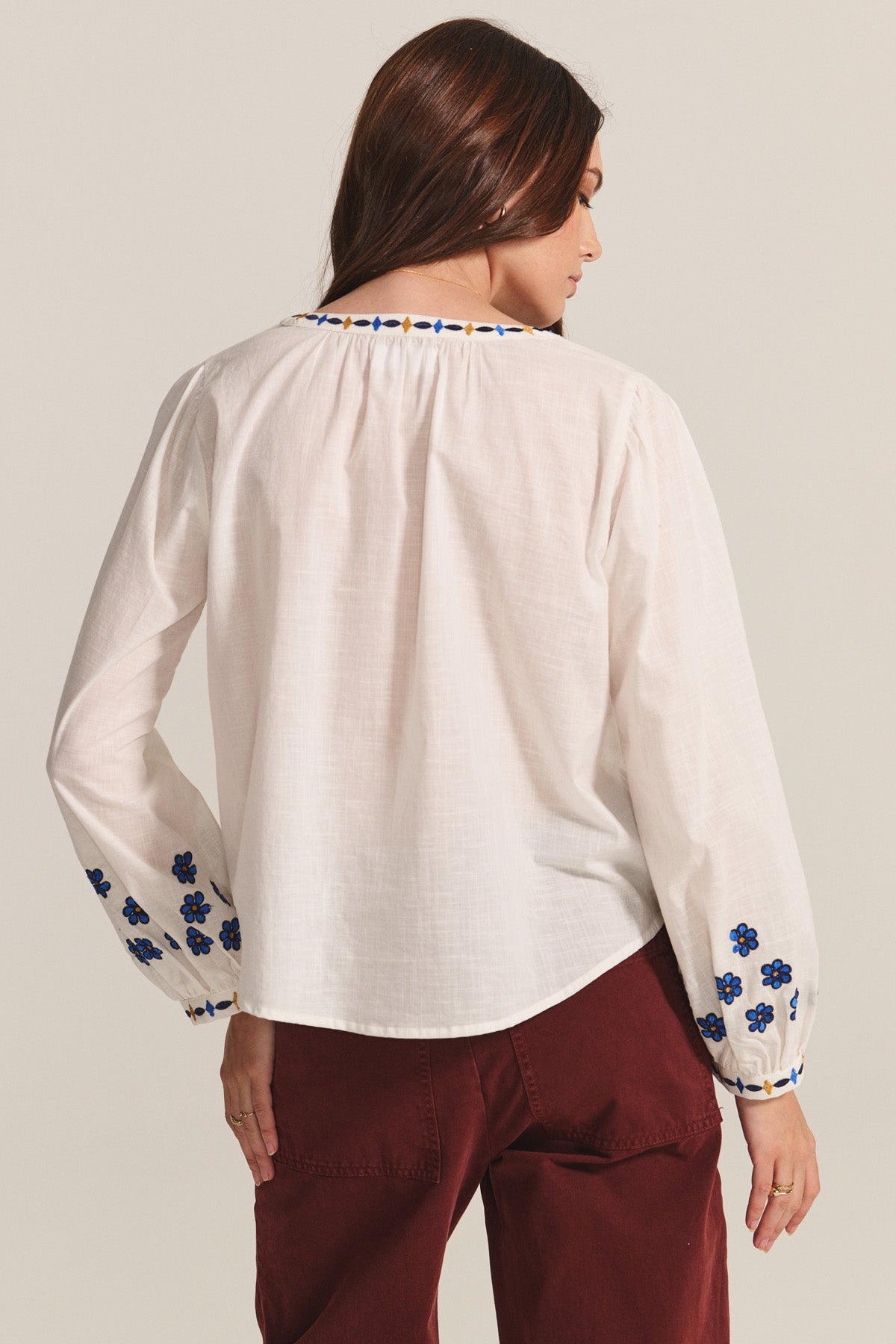 A woman with long brown hair, seen from behind, wears the AMANI BLOUSE by Velvet by Graham & Spencer—a white cotton top with blue floral embroidery—paired with maroon pants.