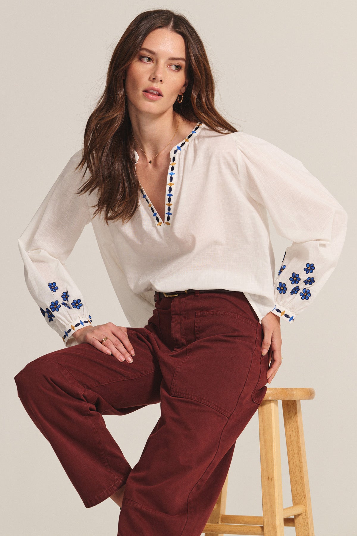 A woman sits on a wooden stool against a plain background, wearing the AMANI BLOUSE by Velvet by Graham & Spencer—white with blue embroidery—paired with maroon pants.-40672276873409