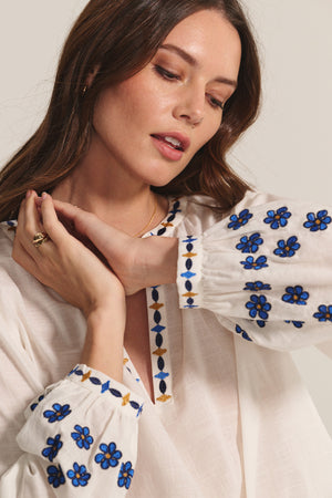 A woman models the AMANI BLOUSE by Velvet by Graham & Spencer, featuring a flowy white design with blue and yellow floral embroidery, posing with her hands raised near her face.