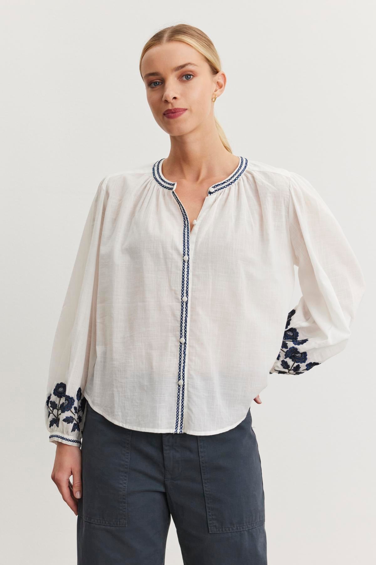   Someone wearing the RAHWA TOP by Velvet by Graham & Spencer, featuring a white blouse with floral embroidery on the sleeves, paired with dark pants, stands against a plain background. 