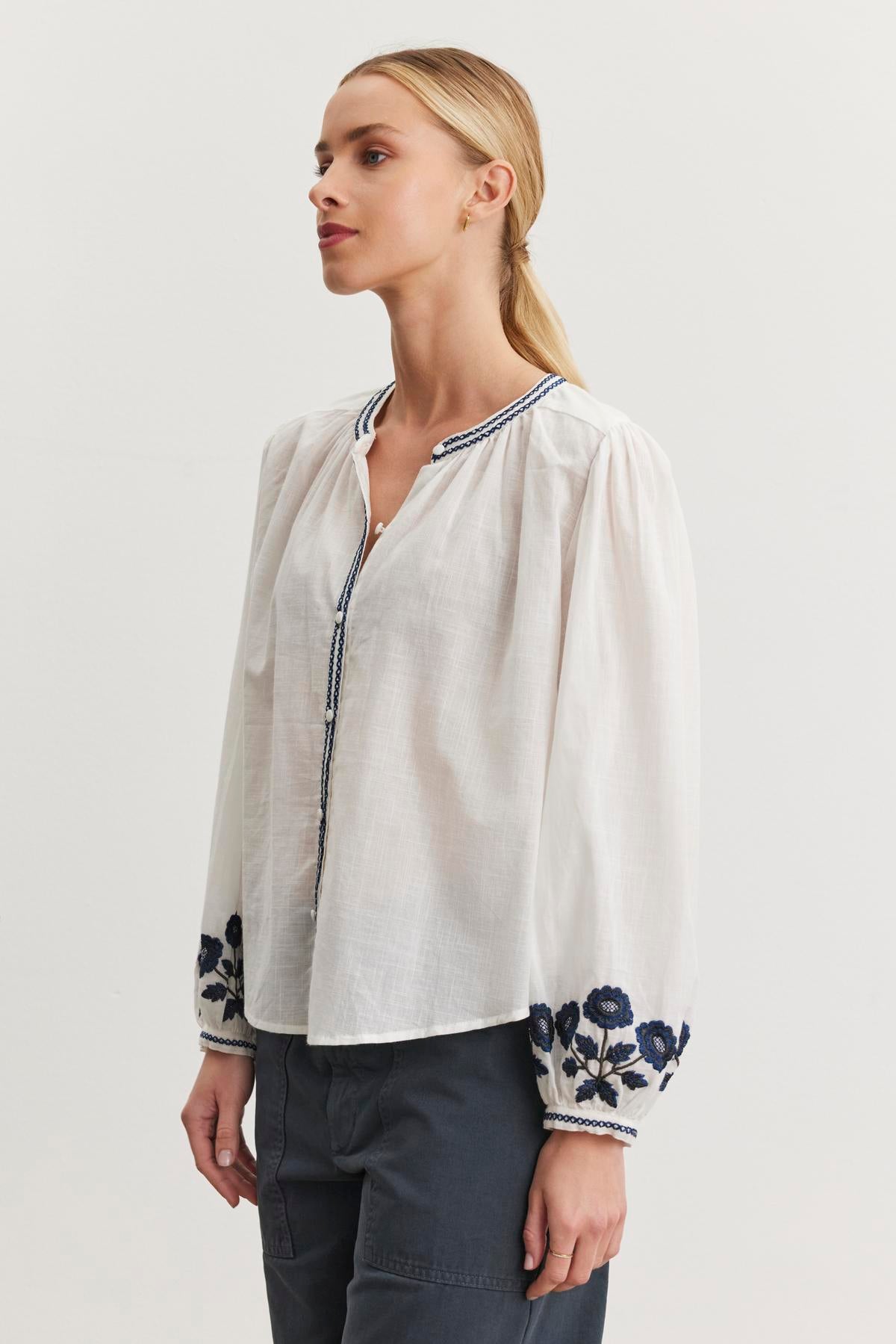   A woman poses against a plain backdrop, wearing the Velvet by Graham & Spencer RAHWA TOP— a lightweight cotton top with blue floral embroidery on the sleeves—paired with dark pants. 