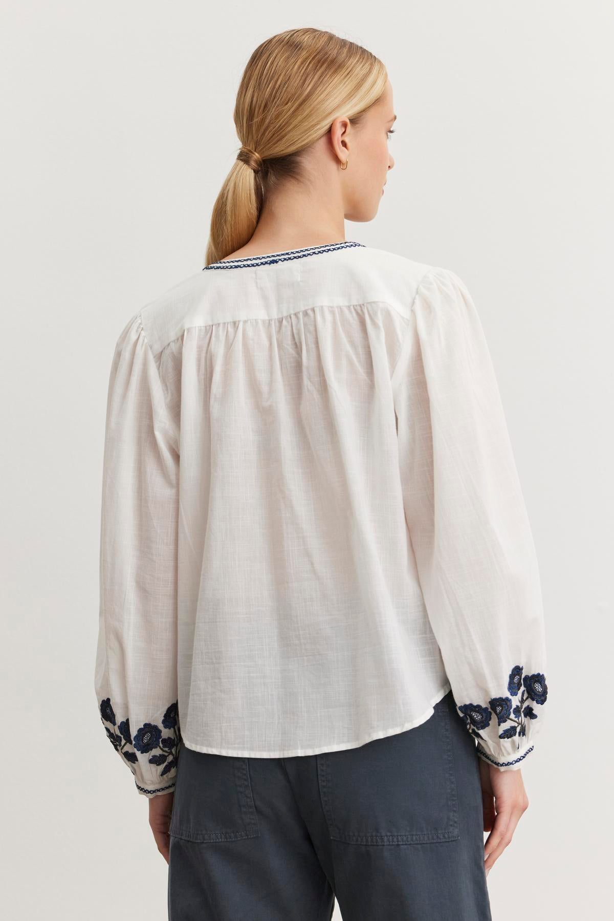   A woman with blond hair in a ponytail wears the RAHWA TOP, featuring blue floral embroidery by Velvet by Graham & Spencer, seen from behind. 