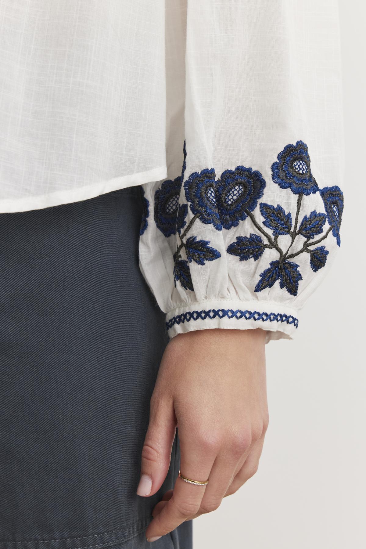   Close-up of a person wearing the RAHWA TOP by Velvet by Graham & Spencer, a lightweight cotton button-front top with blue floral sleeve embroidery, paired with a dark skirt. 