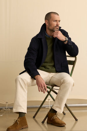 A man with a short beard sits on a green metal folding chair wearing the EMERSON CHORE JACKET by Velvet by Graham & Spencer, paired with a green sweater, white pants, and brown suede shoes against a beige background—ideal for stylish everyday wear.