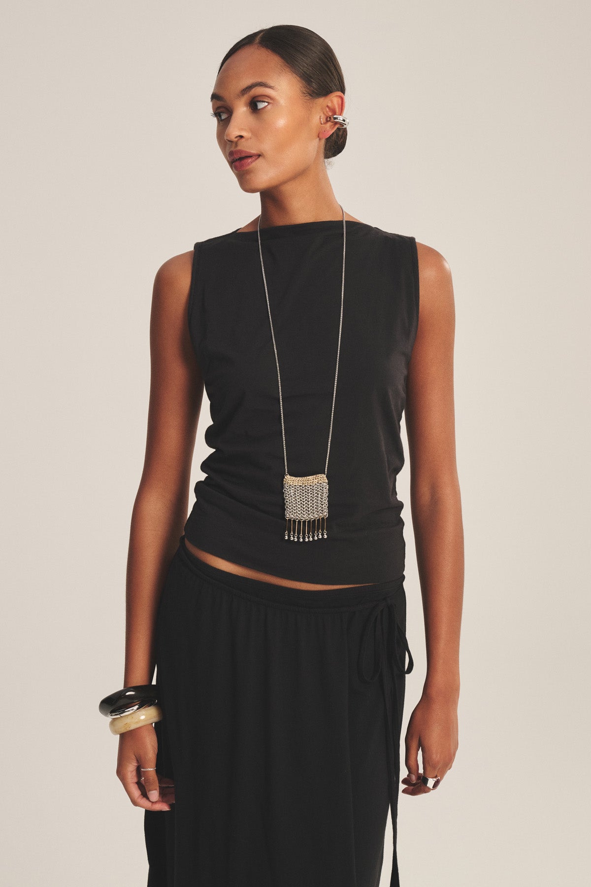 A woman in the EMILIA TANK TOP by Velvet by Graham & Spencer with a skirt, long necklace, and chunky bracelets stands against a plain background, gazing to the side.