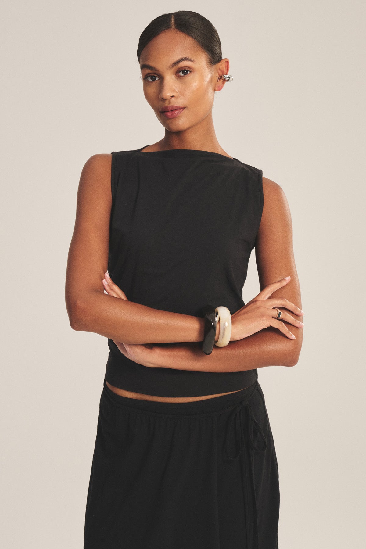 A woman stands with arms crossed against a neutral background, wearing the EMILIA TANK TOP by Velvet by Graham & Spencer styled with a skirt, statement bracelets, and earrings.-41128077033665