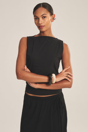 A woman stands with arms crossed against a neutral background, wearing the EMILIA TANK TOP by Velvet by Graham & Spencer styled with a skirt, statement bracelets, and earrings.