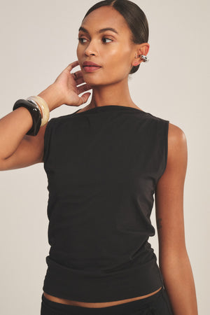 A woman poses against a neutral background wearing the EMILIA TANK TOP by Velvet by Graham & Spencer, styled sleeveless and cropped, with large bracelets and an ear cuff, one hand touching her neck.