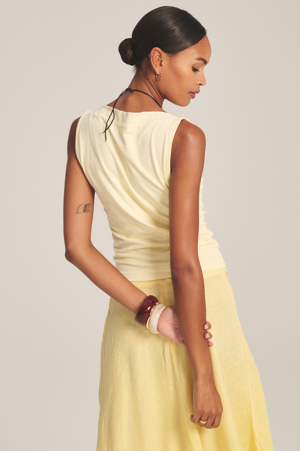Woman stands with her back to the camera, wearing the EMILIA TANK TOP in cream by Velvet by Graham & Spencer and a yellow skirt. Her hair is styled in a low bun, and her left hand rests behind her back.-41128076738753