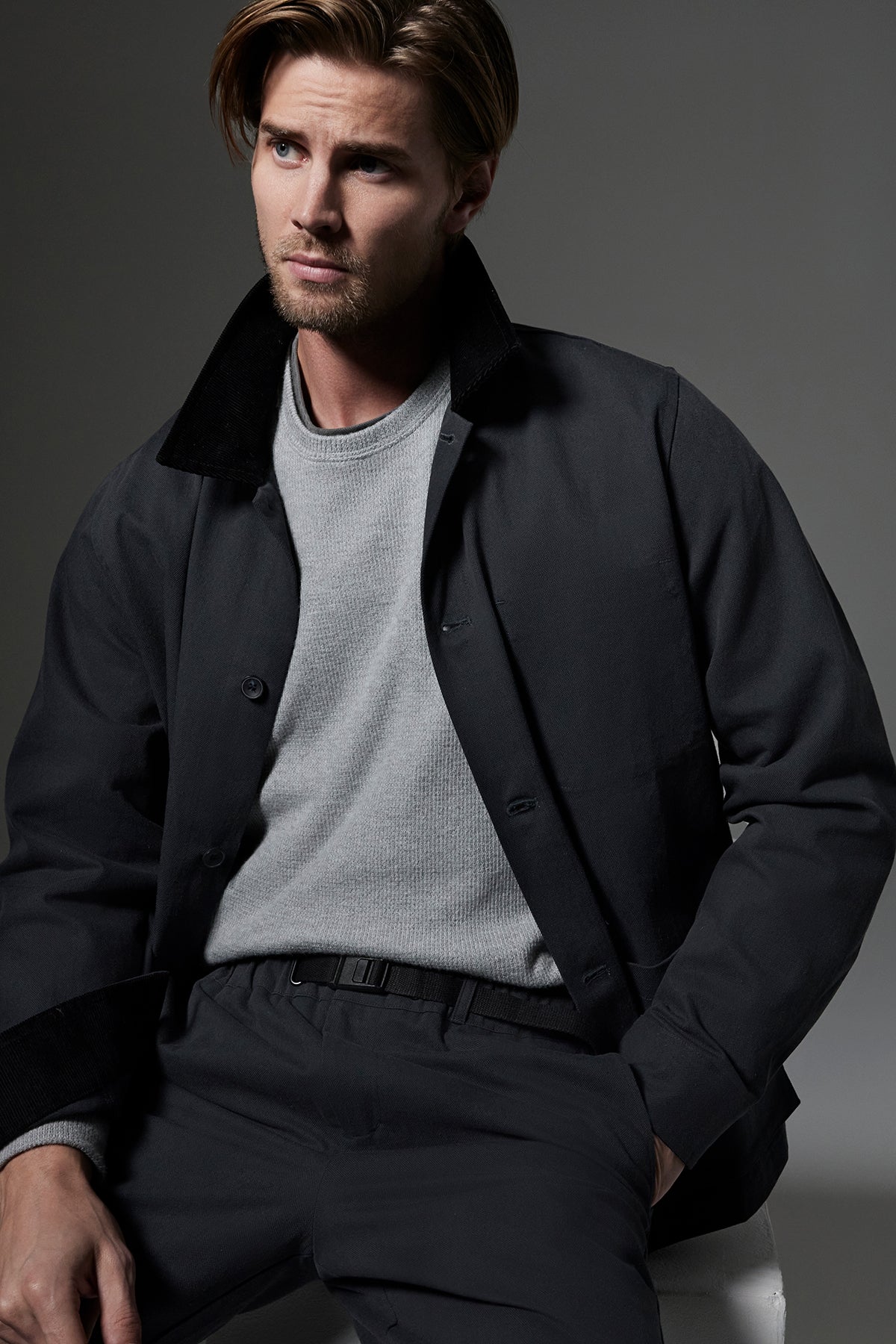   A man with light brown hair and a beard is wearing a grey sweater and the EMORY JACKET by Velvet by Graham & Spencer. He is seated against a neutral backdrop, looking to the side with a serious expression. 