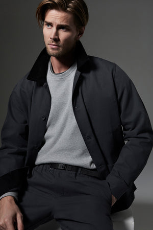 A man with light brown hair and a beard is wearing a grey sweater and the EMORY JACKET by Velvet by Graham & Spencer. He is seated against a neutral backdrop, looking to the side with a serious expression.