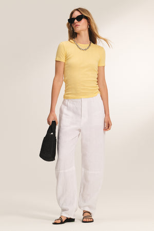 A woman stands against a neutral background wearing the ENATI POINTELLE TEE by Velvet by Graham & Spencer in yellow, styled with white wide-leg pants, black sandals, sunglasses, a chain necklace, and carrying a black bag.