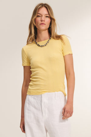 A woman stands against a plain background wearing the ENATI POINTELLE TEE by Velvet by Graham & Spencer—a short-sleeve yellow fitted top with ribbed pointelle details—paired with white pants and a beaded necklace.