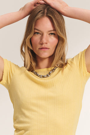 A woman with blonde hair wears the ENATI POINTELLE TEE by Velvet by Graham & Spencer—a yellow ribbed fitted tee—paired with a chunky silver necklace, standing with her arms raised above her head.