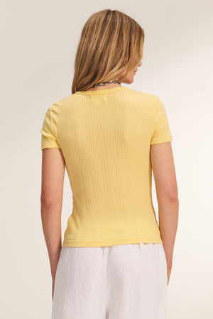 A person with shoulder-length blonde hair, facing away, wears the ENATI POINTELLE TEE by Velvet by Graham & Spencer—a short-sleeved, yellow ribbed top—paired with white pants.