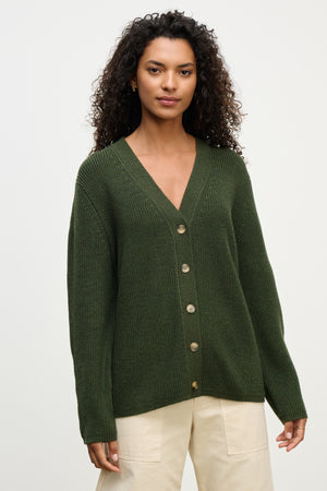 A woman with long curly hair wears the MONET CARDIGAN by Velvet by Graham & Spencer—a dark green, loose fit wool blend V-neck style—paired with beige pants, standing against a plain light background.