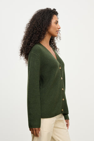 A woman with curly hair models the MONET CARDIGAN by Velvet by Graham & Spencer—a dark green, loose fit wool blend V-neck—paired with light beige pants, standing in profile against a plain white background.