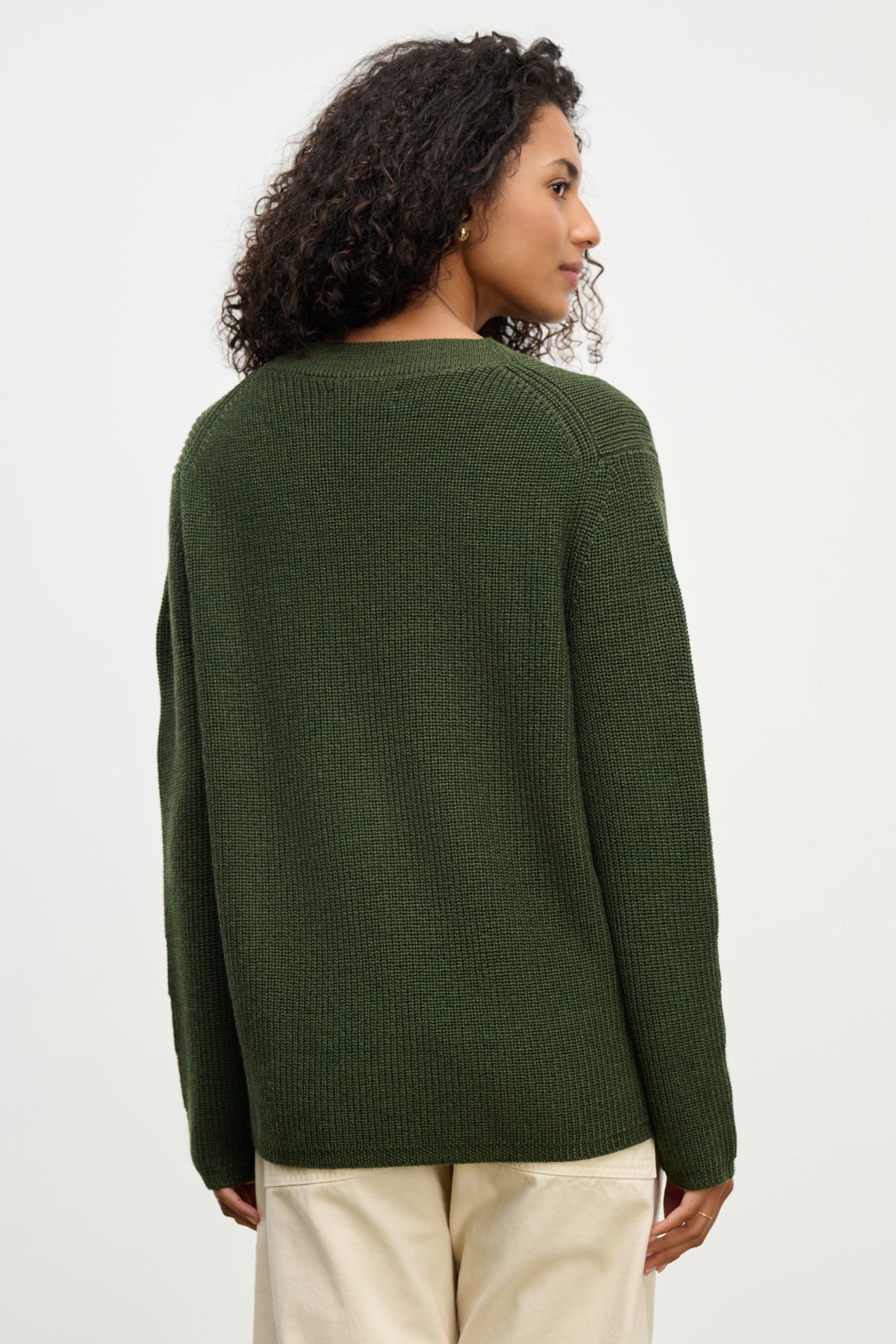 A person stands with their back to the camera, wearing the MONET CARDIGAN by Velvet by Graham & Spencer—a dark green, loose fit wool blend—paired with light-colored pants against a plain background.