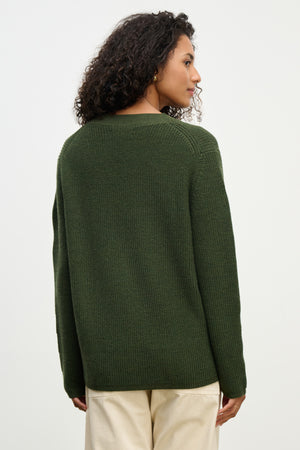 A person stands with their back to the camera, wearing the MONET CARDIGAN by Velvet by Graham & Spencer—a dark green, loose fit wool blend—paired with light-colored pants against a plain background.