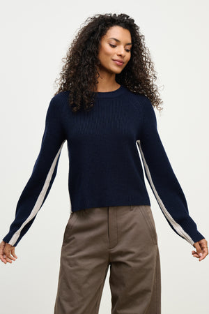 A woman models the SAVANNAH SWEATER by Velvet by Graham & Spencer, a navy wool-blend with contrast sleeve stripes and a crew neck, styled with brown high-waisted pants against a light plain background.