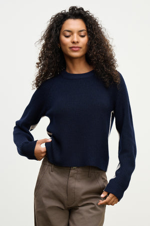 A person with curly hair wears the SAVANNAH SWEATER by Velvet by Graham & Spencer—navy blue, wool blend, crew neckline—paired with taupe pants, standing against a plain light background.