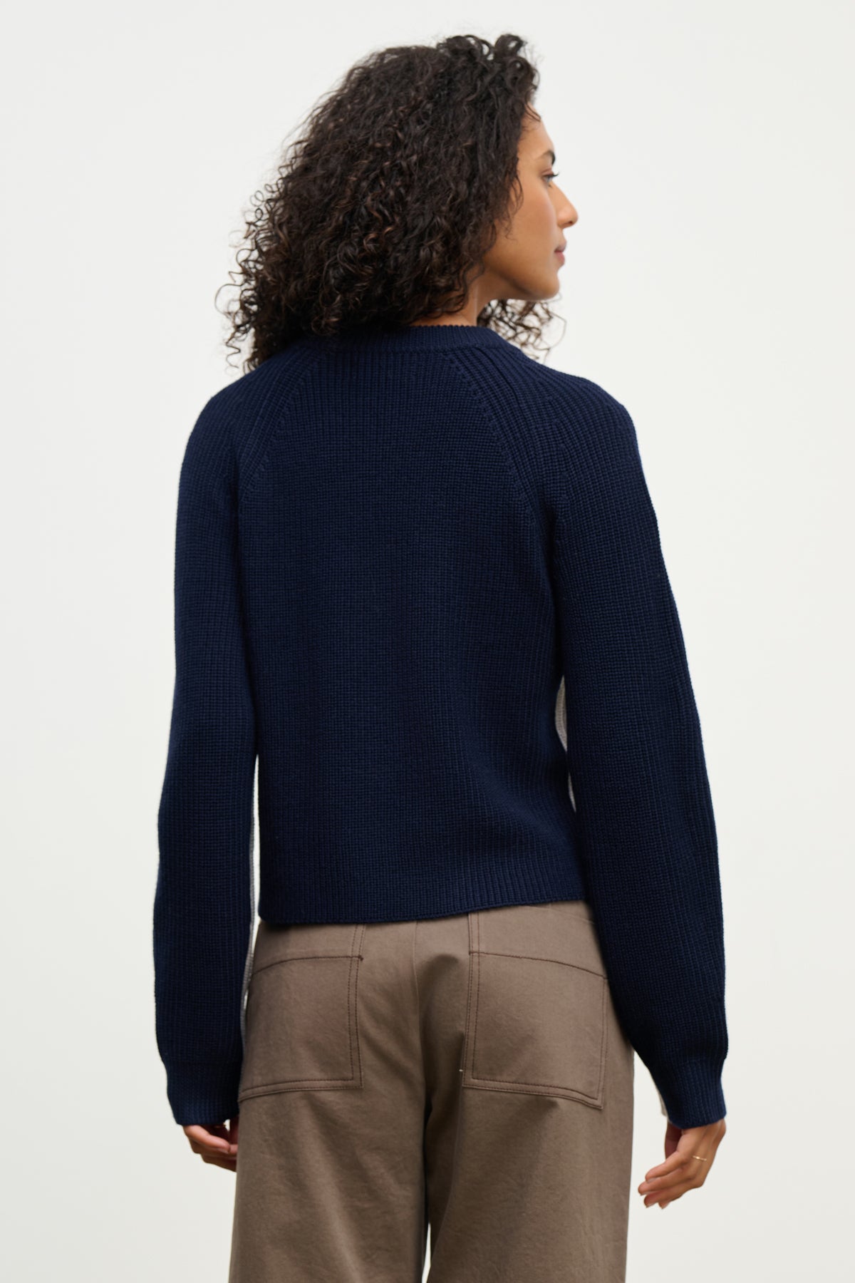   A person with curly hair, shown from the back against a plain light background, wears the SAVANNAH SWEATER by Velvet by Graham & Spencer—a navy blue wool blend crewneck—paired with taupe pants. 