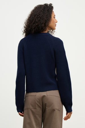 A person with curly hair, shown from the back against a plain light background, wears the SAVANNAH SWEATER by Velvet by Graham & Spencer—a navy blue wool blend crewneck—paired with taupe pants.