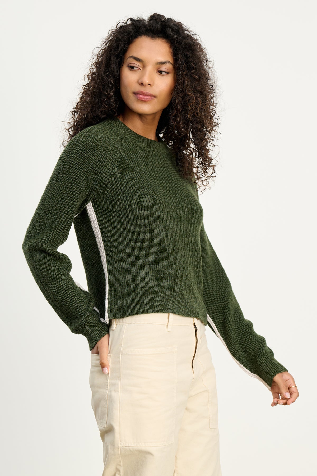   A person with curly hair wears the SAVANNAH SWEATER by Velvet by Graham & Spencer in dark green, paired with light-colored pants, standing against a plain white background. 