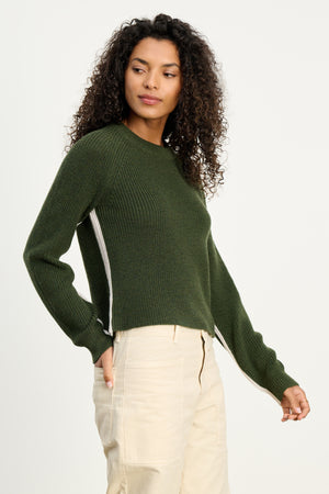 A person with curly hair wears the SAVANNAH SWEATER by Velvet by Graham & Spencer in dark green, paired with light-colored pants, standing against a plain white background.