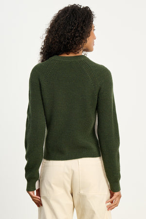 A person with curly hair is shown from the back, wearing the SAVANNAH SWEATER by Velvet by Graham & Spencer—a dark green wool blend crewneck—paired with light pants against a plain background.