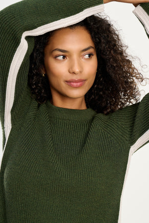 A woman with curly hair wears the SAVANNAH SWEATER by Velvet by Graham & Spencer, featuring green wool blend fabric, contrast stripes, and a crew neckline. She raises her arms above her head, glancing sideways against a plain backdrop.
